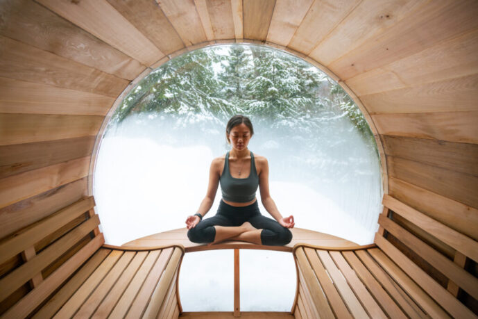 A woman practices yoga in a wooden barrel sauna with a panoramic window showing the forest in the Laurentian Mountains.